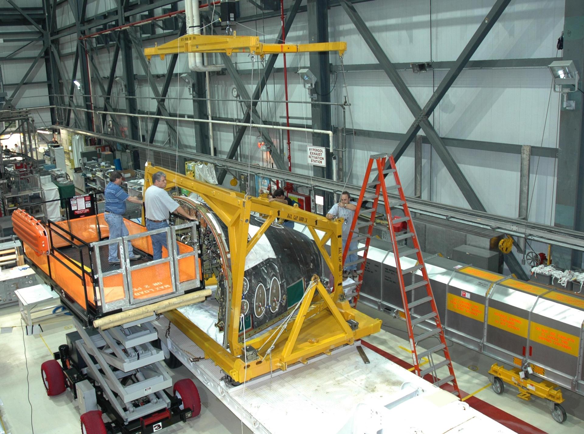 Das Bug-RCS-Modul des Space Shuttle Atlantis hängt in einer gelben Transportvorrichtung in einer Werkhalle, daneben stehen Techniker