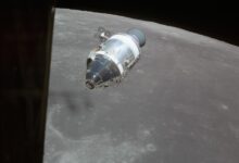 Das Apollo-15-Raumschiff im Mondorbit vor der grauen Mondoberflaeche, an der Flanke des Servicemoduls sind zwei kreuzfoermige Triebwerksgruppen zu erkennen