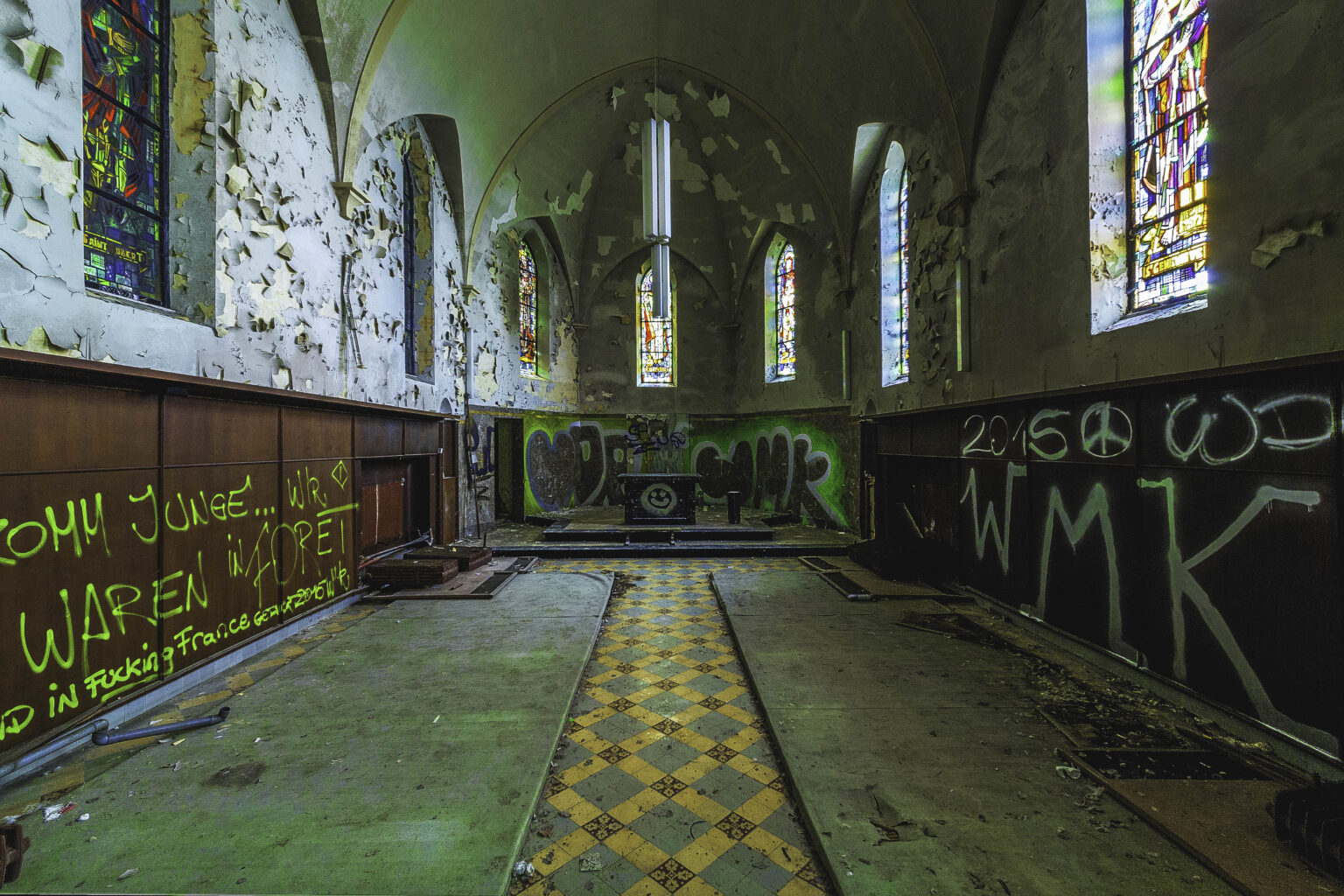 Kapellenchor mit Buntglasfenstern, Holzvertäfelung und gelbem Schachbrettboden im Lost Place