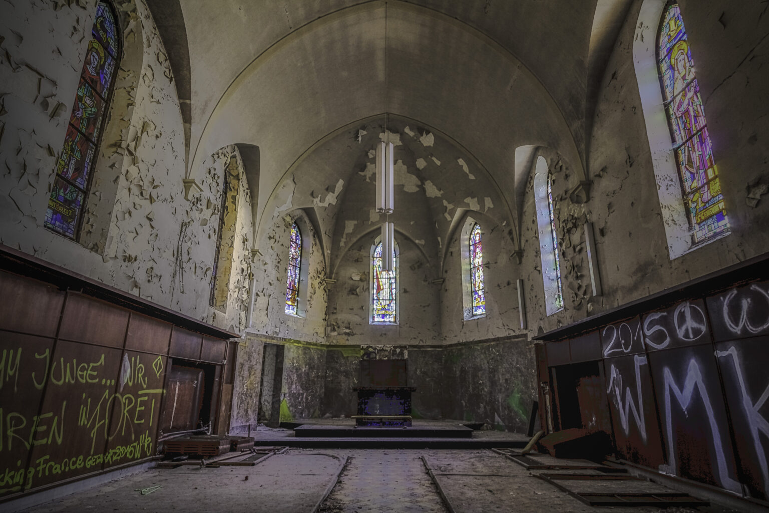 Weiter Kapellenchor mit Lanzett-Buntglasfenstern und abblätternden Wänden am Altar im Lost Place