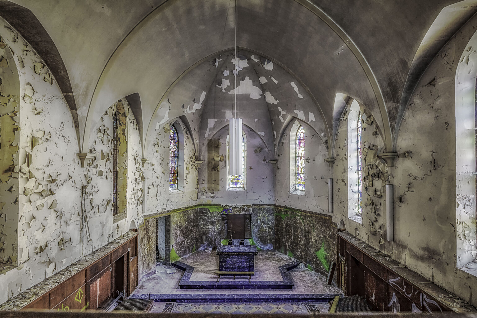 Kapellenchor von der Empore mit Gewölbeapsis, Buntglas und moosigem Altarpodest im Lost Place