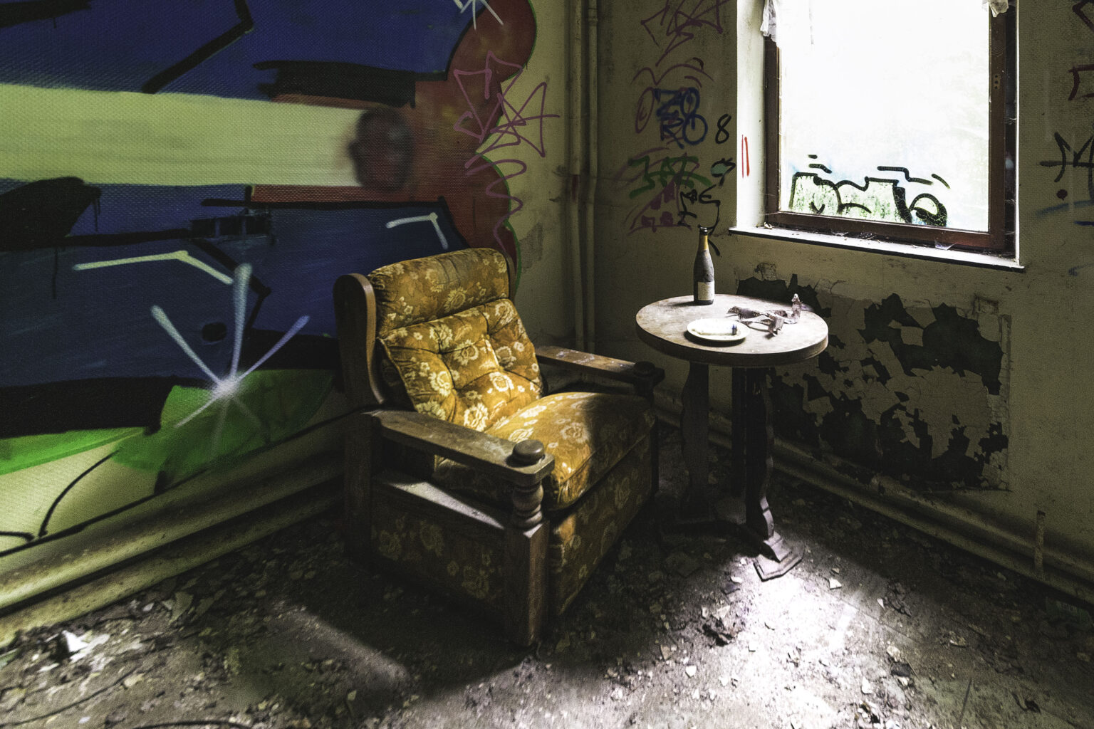 Gelber Polstersessel mit Beistelltisch vor graffitibedeckter Wand am Fenster im Lost Place