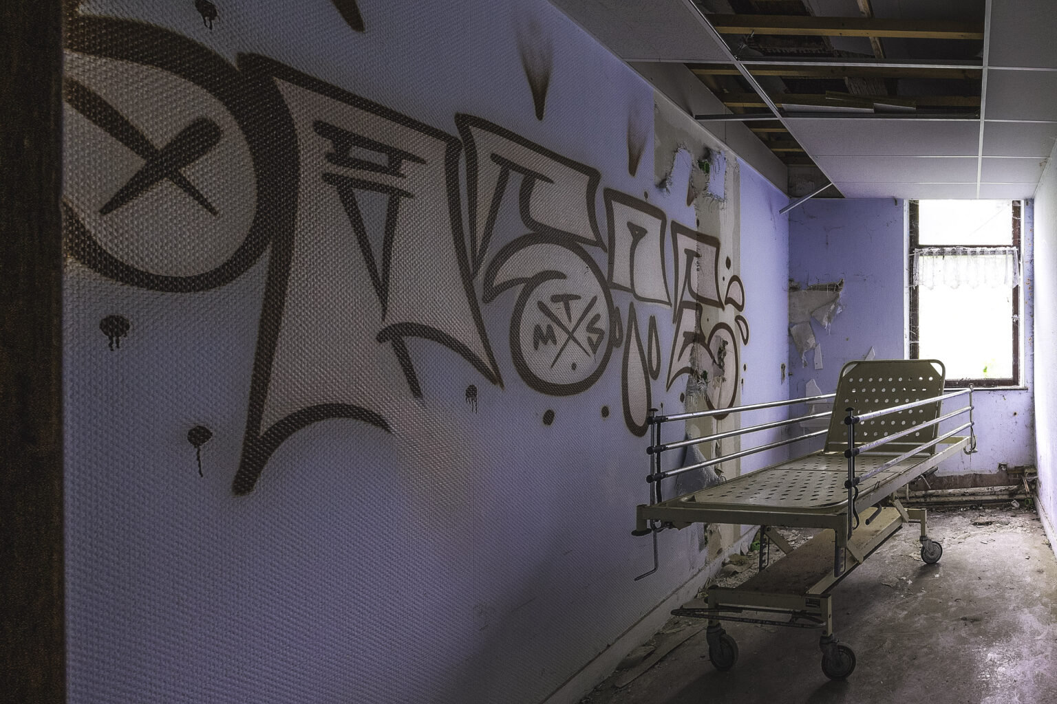 Korridor mit altem Krankenbett vor einer graffitibedeckten Wand am Fenster im Lost Place