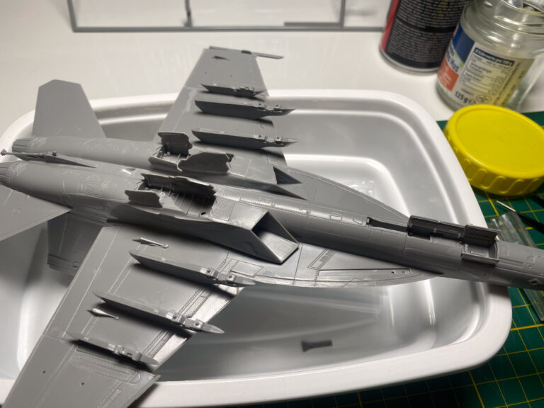 Unterseite eines grauen F/A-18-Modells mit Bewaffnung auf weißer Schale