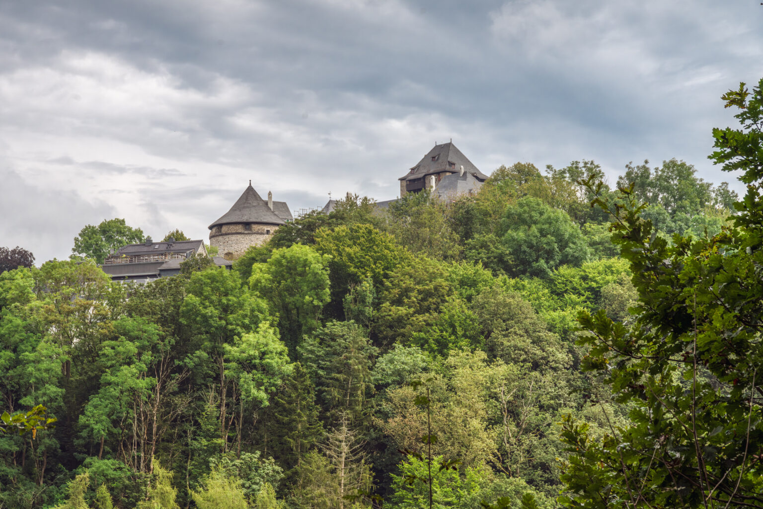 Schloss mit Turm auf bewaldetem Höhenzug unter wolkigem Himmel im Bergischen Land