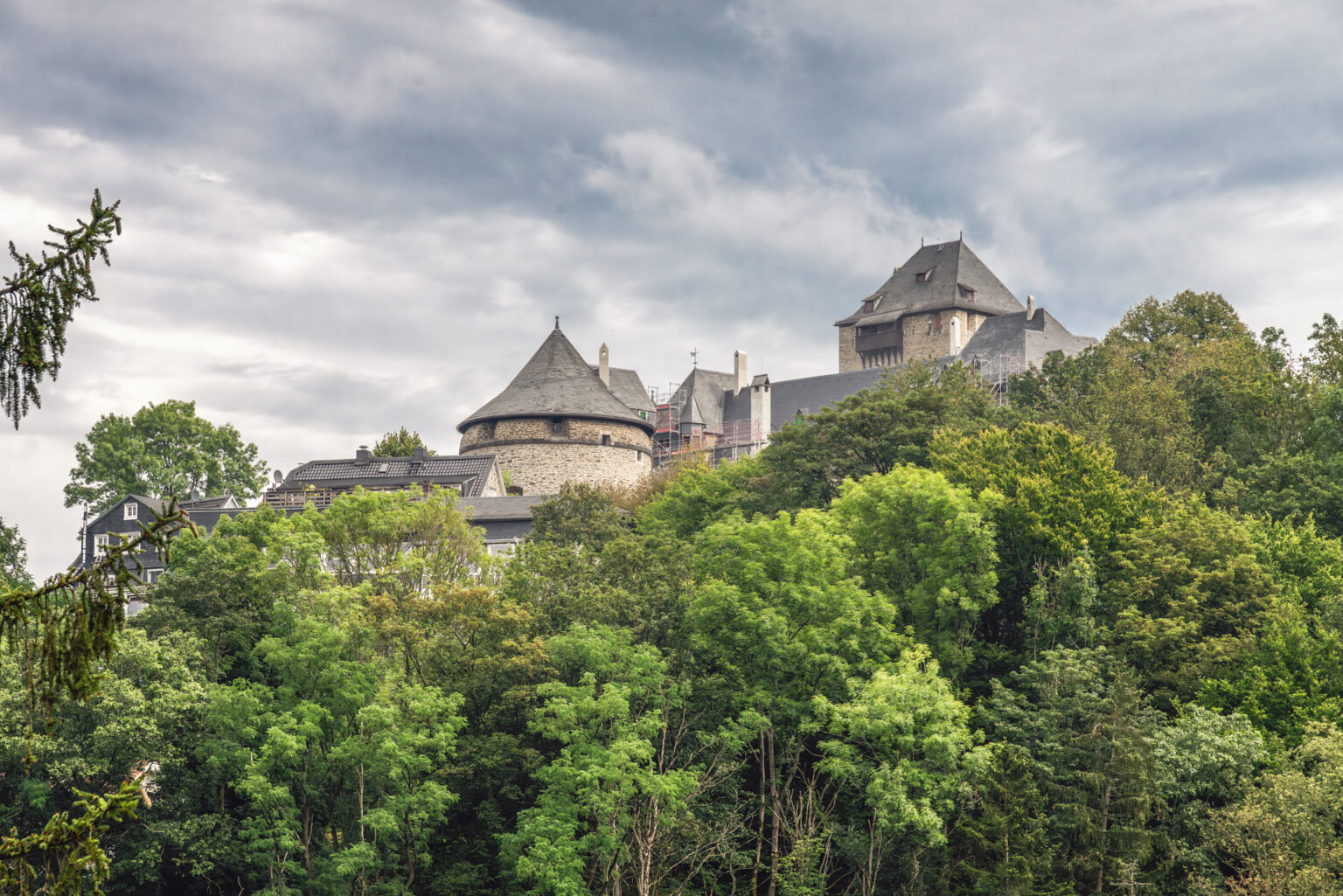 Schloss mit Rundturm auf bewaldetem Hügel unter Wolkenhimmel im Bergischen Land