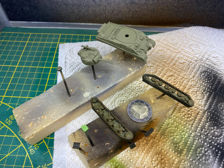 Olivgrün lackierte Turm- und Wannenteile zweier Panzermodelle auf Stützen mit Zwei-Euro-Münze als Größenvergleich