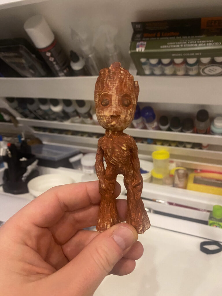 Braun bemalte Baby-Groot-Figur aus dem 3D-Druck in der Hand vor einem Farbregal