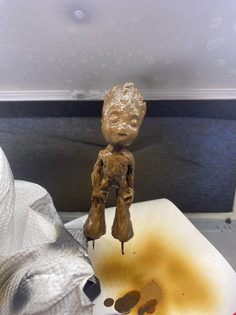 Braun lasierte Baby-Groot-Figur mit feuchter Farbe auf einem Haltestift in der Lackierbox