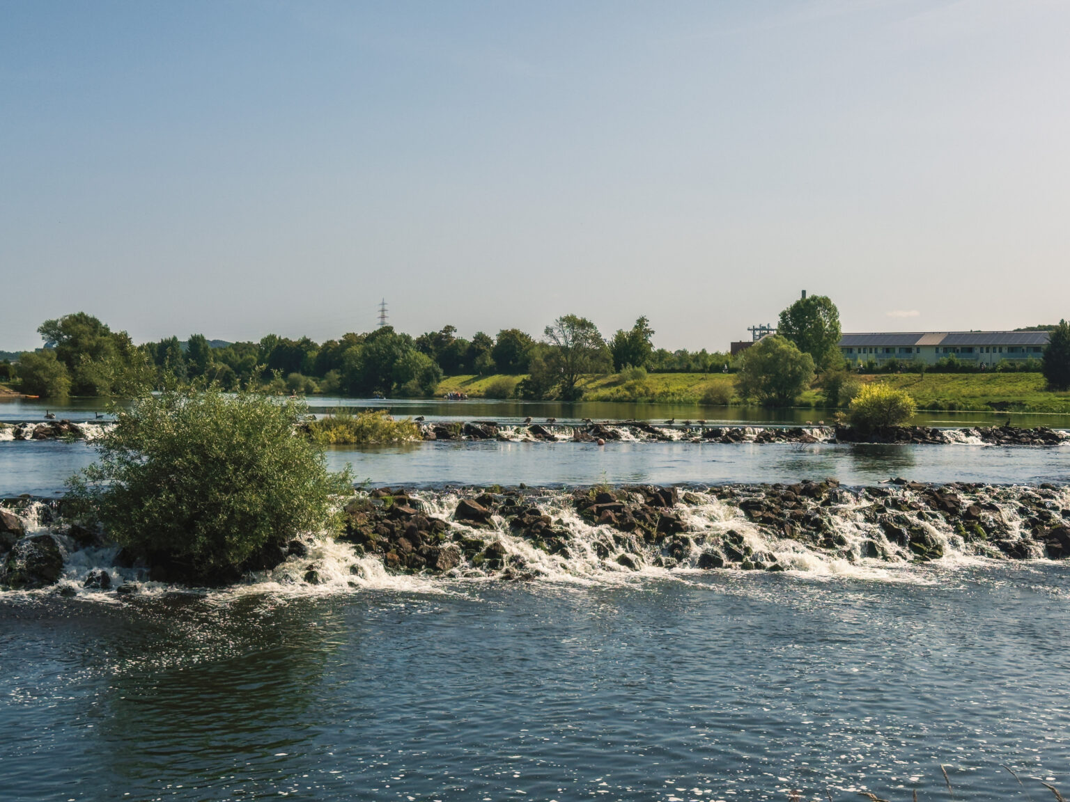 Flusswehr der Ruhr mit Felsschwelle und überlaufendem Wasser vor einem Gewerbebau