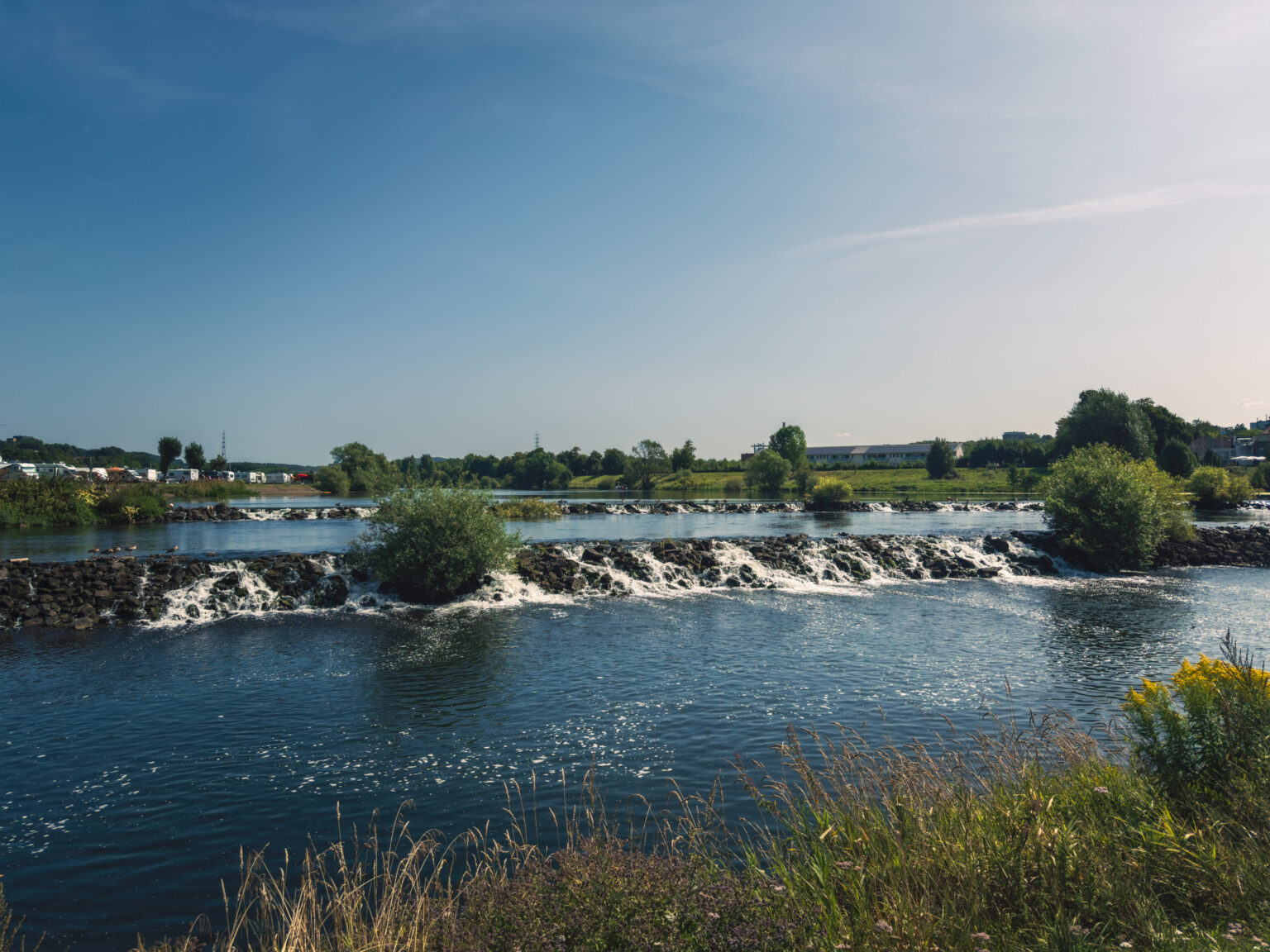 Flusswehr mit überlaufendem Wasser an der Ruhr, im Hintergrund ein Campingplatz