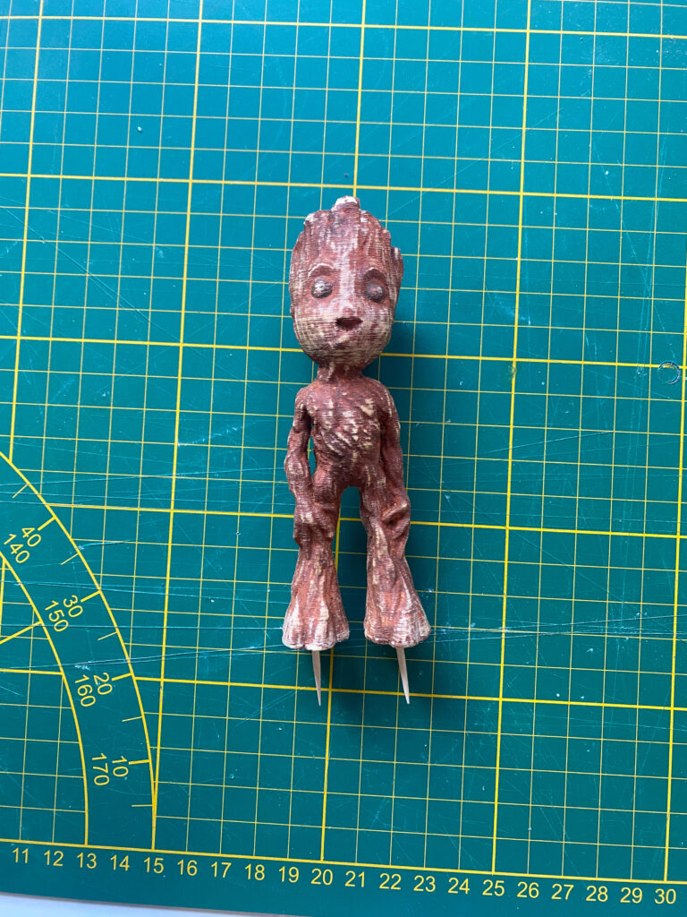 Braune Baby-Groot-Figur auf grüner Schneidematte mit Zahnstochern an den Füßen