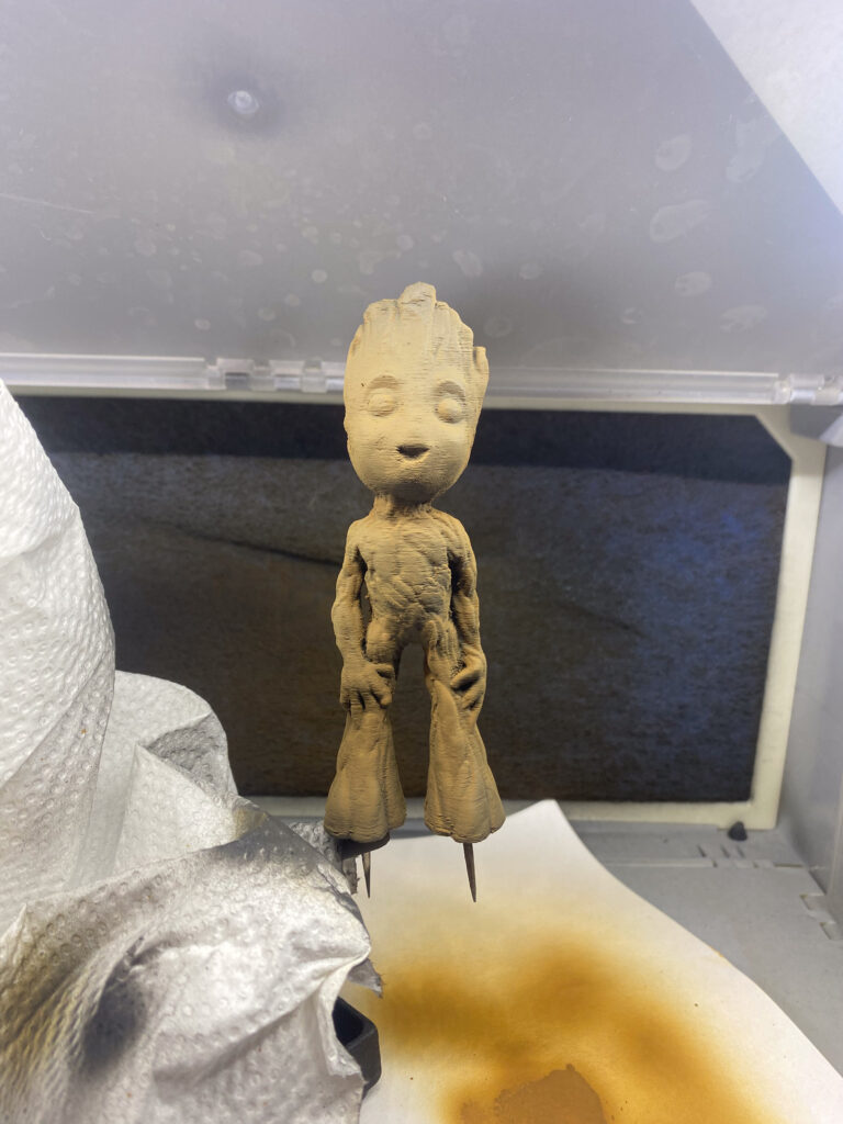 Geschliffene holzfarbene Baby-Groot-Figur auf einem Haltestift in der Lackierbox