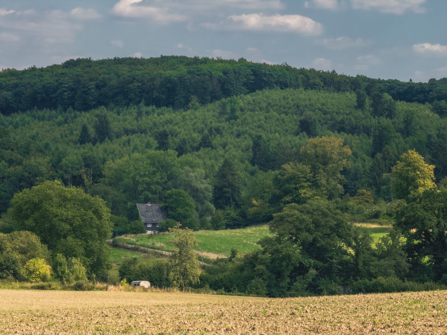 Bewaldeter Hügel mit einem einzelnen Haus über einem abgeernteten Feld im Bergischen Land