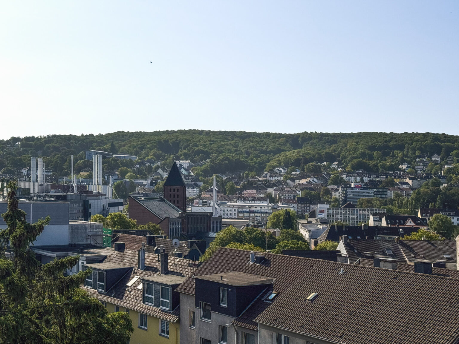 Stadtpanorama mit Kirchturm und Industrieschornsteinen vor bewaldeten Höhen