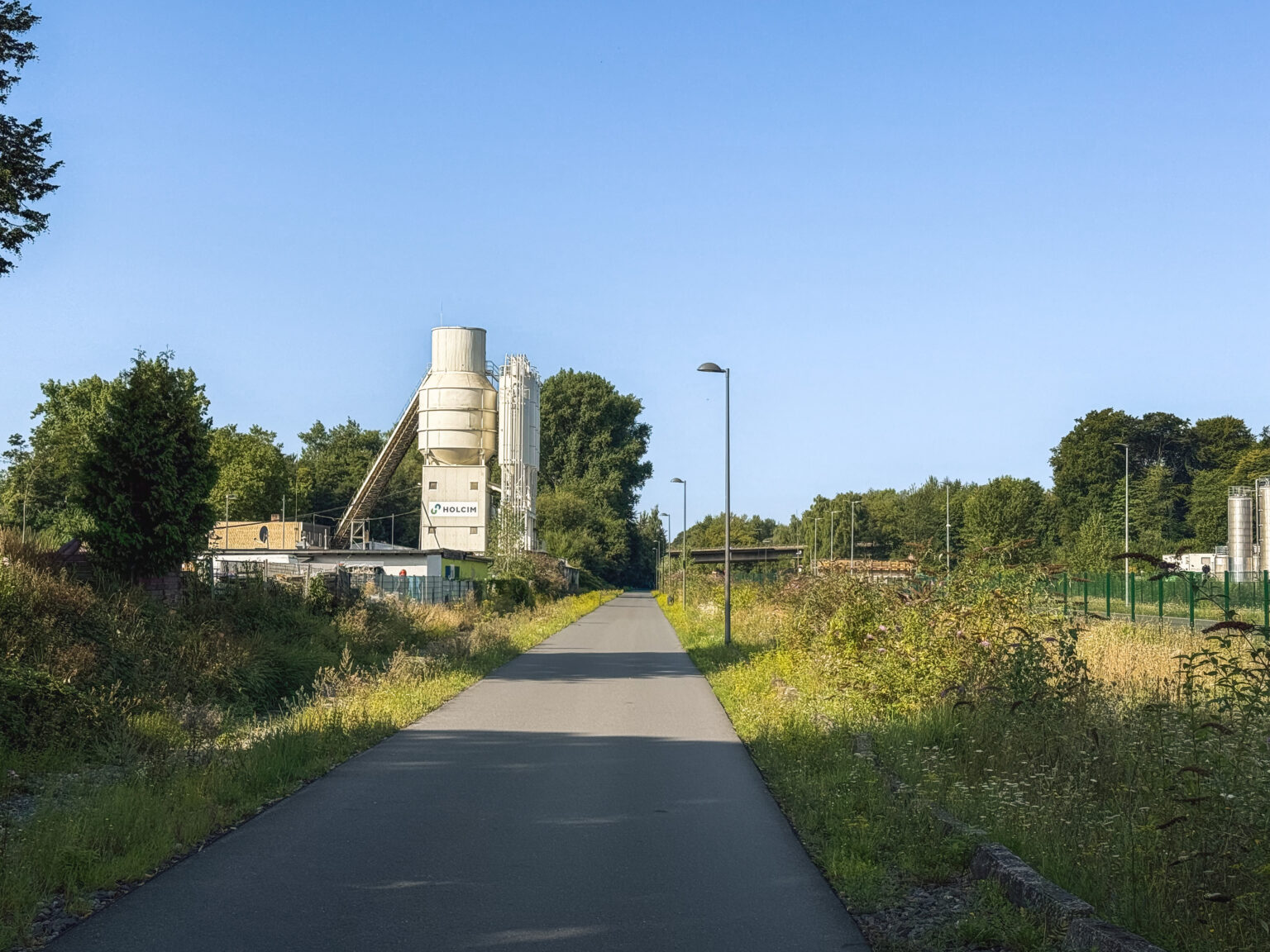 Asphaltierter Radweg durch Sommerwiesen mit einem Zementwerk-Silo im Hintergrund