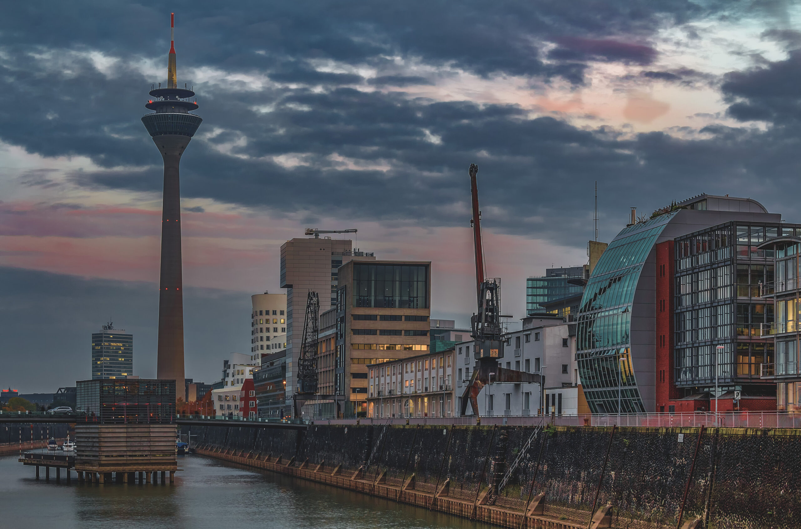 The Rheinturm above the Medienhafen at dusk, old harbour cranes and a curved glass front on the right