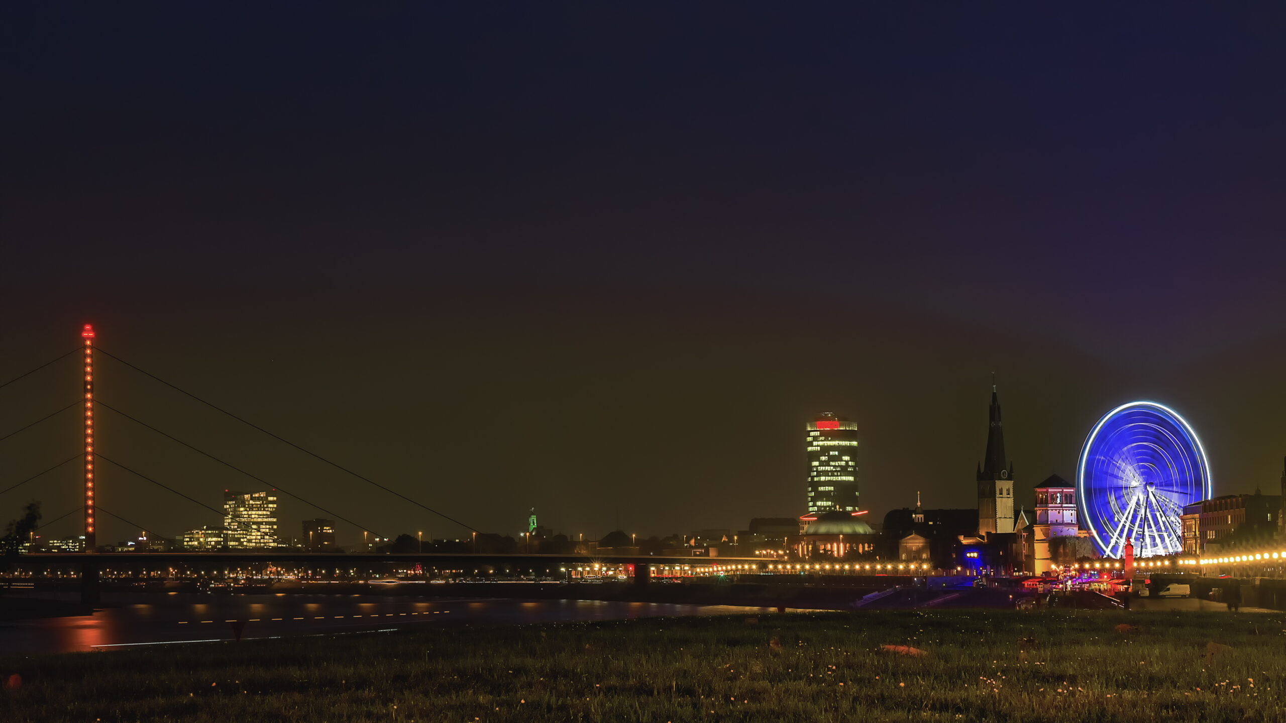 Düsseldorf's old town at night from the left bank of the Rhine, the red-lit pylon of a cable-stayed bridge on the left, a Ferris wheel glowing blue on the right