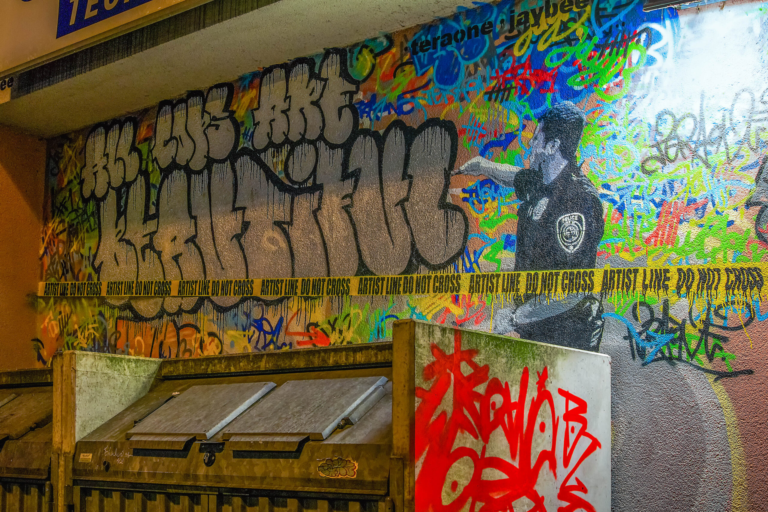 Graffiti wall with a sprayed police officer and big silver lettering, yellow tape reading Artist Line do not cross in front
