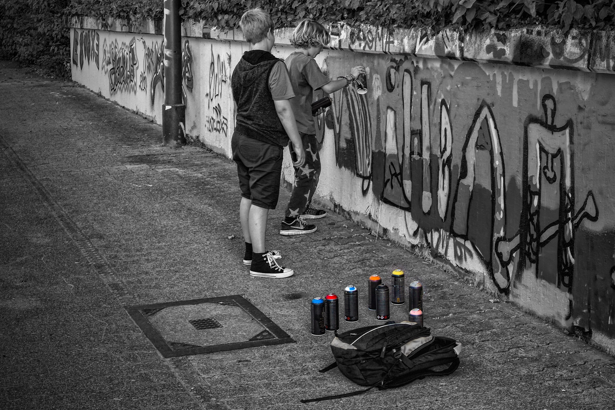 Black-and-white shot of a sprayer at a graffiti wall, spray cans with coloured caps lined up in front of him
