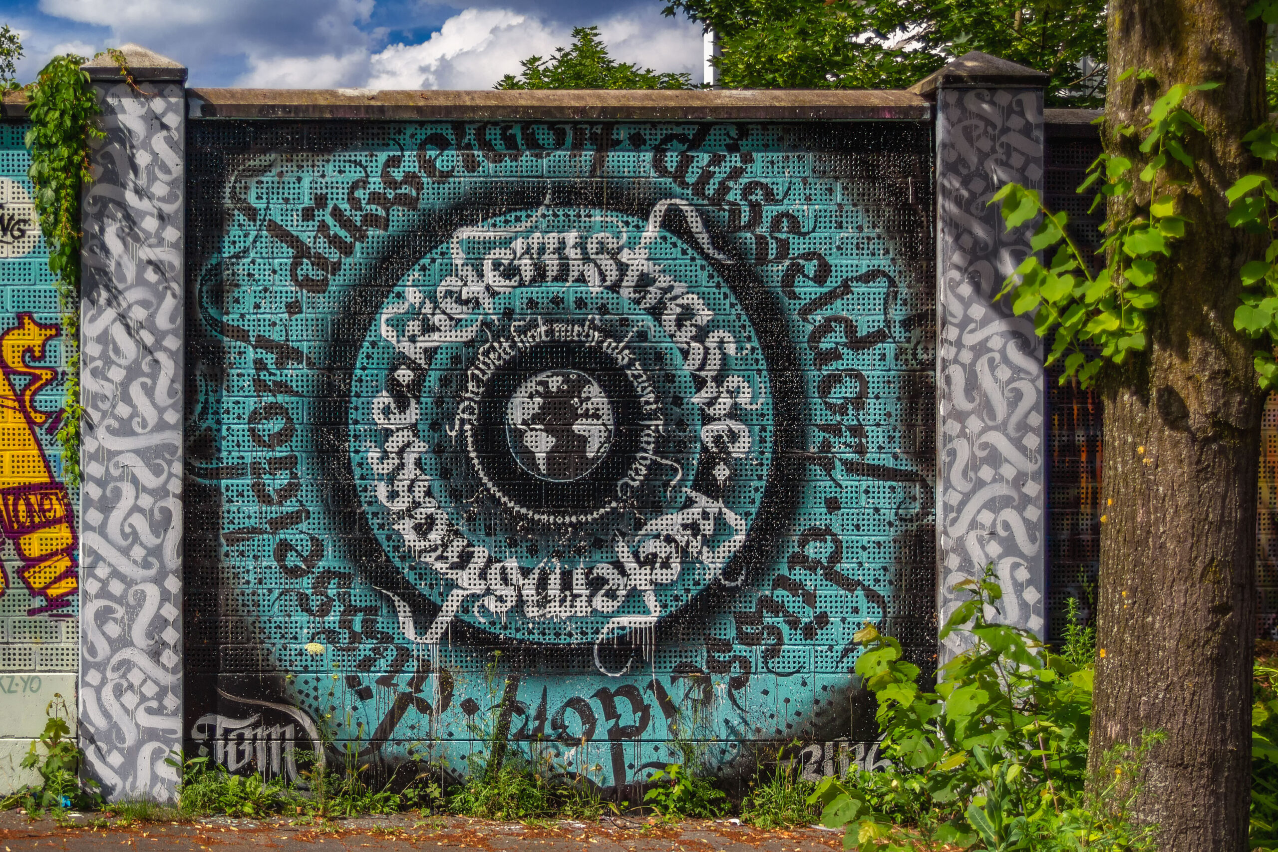 Round turquoise mural with the lettering Kiefernstraße Düsseldorf and a globe in the middle, framed by two pillars